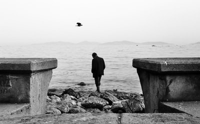 During the day, the man wearing a black jacket standing on the rocks near the water
